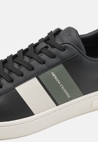Sneaker in pelle nera con superficie texture, caratterizzato da strisce grigie e bianche. Logo "Armani Exchange" stampato di lato, lacci neri, suola in gomma.