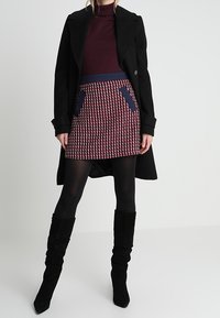 Musta takki viininpunaisen pooloneuleen päällä, yhdistetty kuvioituun punaiseen ja laivastonsiniseen hameeseen, jossa on taskut, sekä mustat polven yläpuolelle ulottuvat saappaat.