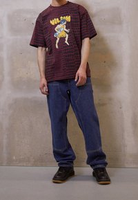 Mand iført en rød og sort mønstret T-shirt med en tegneserie-hulemand der holder et skateboard, blå jeans og sorte sko, står indendørs.