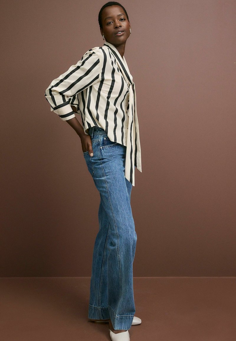 Stribet bluse i creme og sort med knyttet front, parret med lyseblå flare-jeans. Grå sko fuldender outfittet. Brun baggrund.