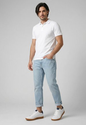 Mann steht, trägt ein weißes Poloshirt, hellblaue hochgekrempelte Jeans und weiße Sneaker mit braunen Sohlen vor einem schlichten grauen Hintergrund.