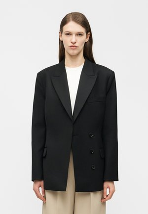 Γυναίκα με ίσια καστανά μαλλιά φορούν υπερμεγέθη μαύρο σακάκι με διπλή σειρά κουμπιών πάνω από λευκό τοπ και μπεζ παντελόνι μπροστά σε απλό φόντο.