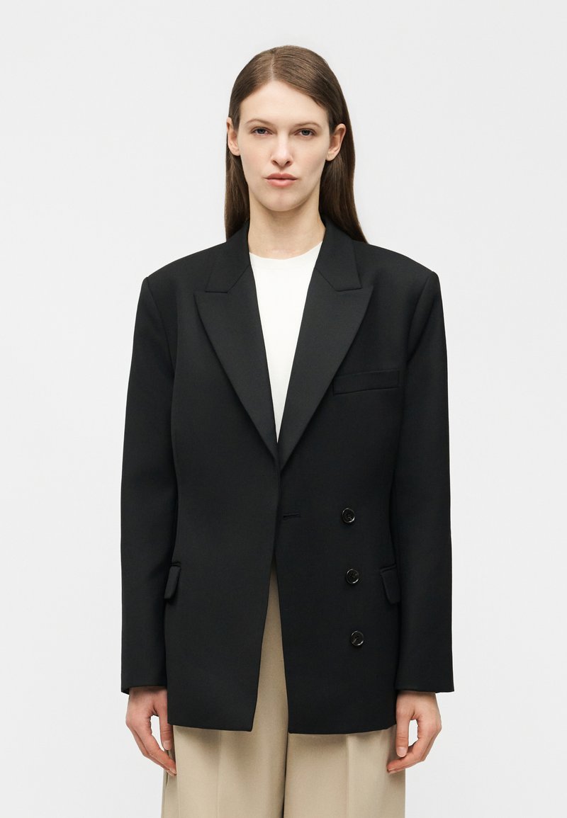 Γυναίκα με ίσια καστανά μαλλιά φορούν υπερμεγέθη μαύρο σακάκι με διπλή σειρά κουμπιών πάνω από λευκό τοπ και μπεζ παντελόνι μπροστά σε απλό φόντο.