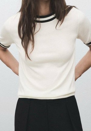 Personne portant une chemise blanche à manches courtes avec un liseré noir au col et aux manches, associée à un pantalon noir taille haute, les mains dans le dos.