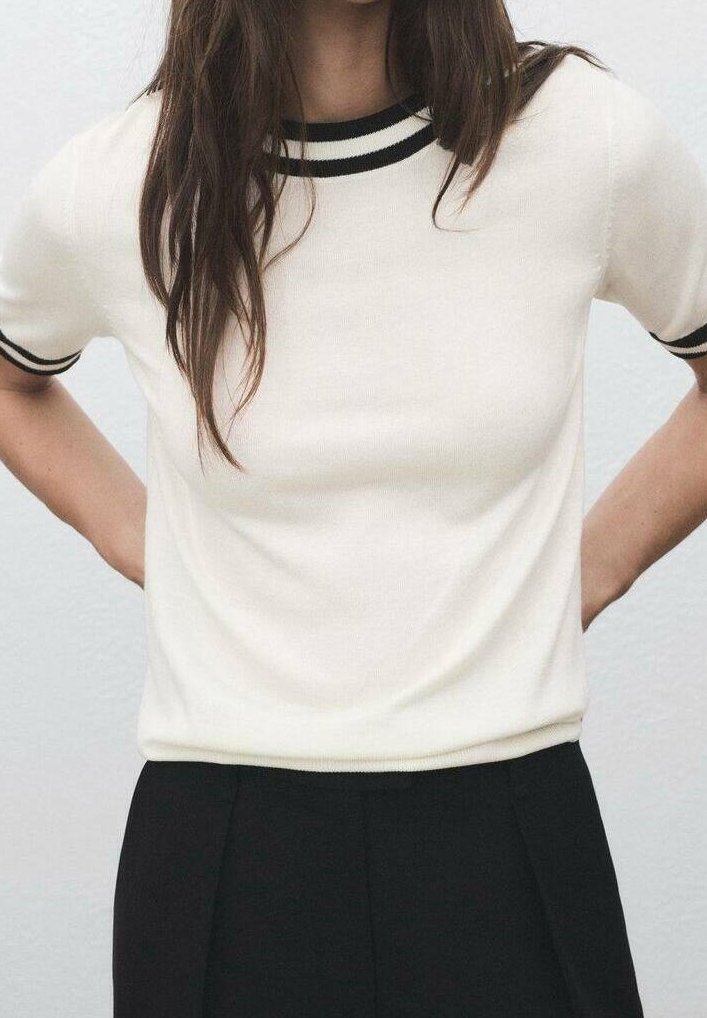 Personne portant une chemise blanche à manches courtes avec un liseré noir au col et aux manches, associée à un pantalon noir taille haute, les mains dans le dos.
