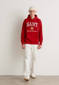 Sudadera roja con letras y logo en blanco, combinada con pantalones blancos y zapatillas beige. El modelo lleva un gorro claro. Look simple y casual.
