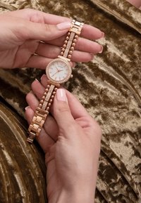 Złoty zegarek na wrist z białą tarczą, ozdobiony kryształami i akcentami w formie kul, na teksturowanym brązowym tle.