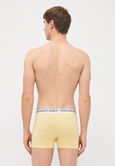 Rumene bombažne boxershorts z belim pasom, na katerem je napis "POLO RALPH LAUREN" v črni barvi. Oseba je prikazana s hrbtne strani.