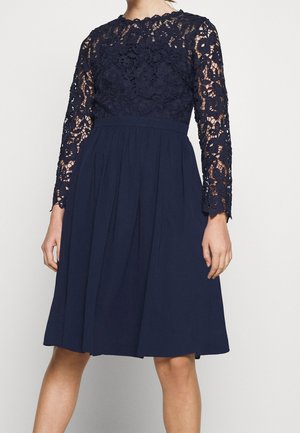 Kobieta ubrana w granatową sukienkę do kolan z długimi rękawami z kwiatowej koronki i jednolitą plisowaną spódnicą, stojąca na jednolitym tle.
