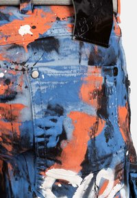 Pantalons en denim bleu avec des éclaboussures de peinture orange, noire et blanche et une patch en cuir noir sur la ceinture.