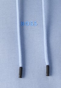 Hanorac bleu deschis din material texturat, cu logo "BOSS" în nuanță de albastru și capete metalice negre pe sforile de strângere.