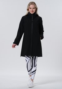 Veste longue noire avec capuche et fermeture éclair à l'avant, associée à des leggings blancs avec des motifs géométriques noirs. Texture lisse et design ajusté.