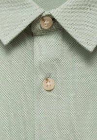 Vihreä polo-paita, jossa on teksturoitu kangas, kaulus ja kaulassa vaalean sävyinen nappi neljässä reiässä.