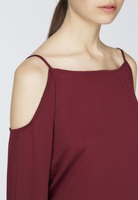 Top bordeaux con spalle scoperte, con spalline sottili, tessuto morbido, vestibilità rilassata e delicate pieghe vicino al collo.