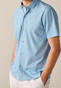 Camisa de manga corta con botones en tejido azul claro con una textura sutilmente rayada, que presenta un cuello clásico y tres botones en la parte delantera.