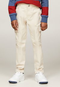 Pantaloni cargo beige con due tasche laterali, in materiale cotone, a taglio slim, abbinati a un maglione a righe rosso e blu e scarpe da ginnastica bianche.