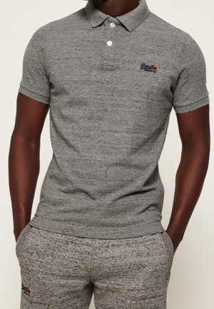 Man draagt een grijs poloshirt met korte mouwen en bijpassende grijze broek, met handen in de zakken, tegen een effen achtergrond.