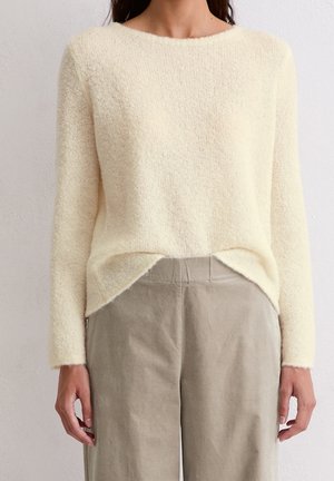 Femme portant un pull duveteux crème à manches longues légèrement rentré dans un pantalon beige taille haute à jambes larges, sur un fond uni.