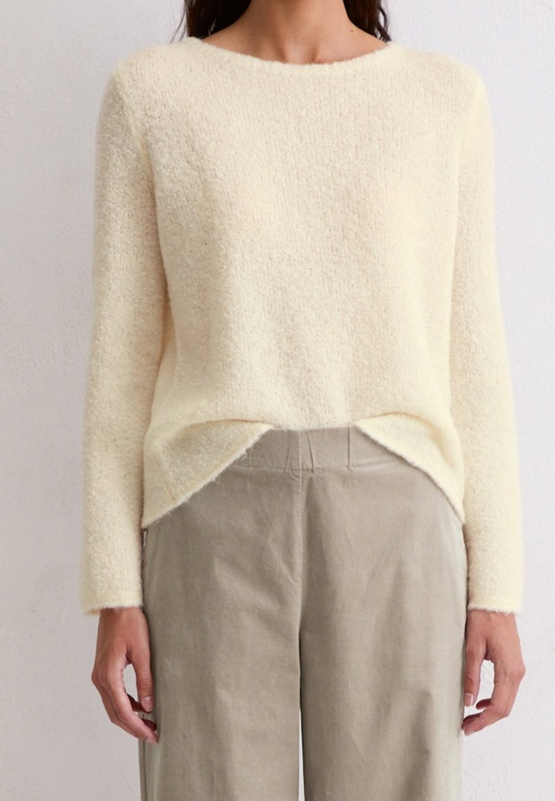 Femme portant un pull duveteux crème à manches longues légèrement rentré dans un pantalon beige taille haute à jambes larges, sur un fond uni.