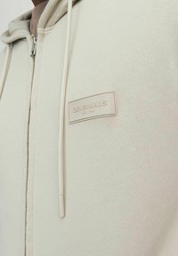 Heledbež zip-up kapuutsärk, millel on tekstuuriga kangas, esiplaanil "ORIGINALS" silt, reguleeritavad nöörid ja sile pind.
