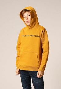 Gele hoodie met een voorzak in de vorm van een kangoeroezak, geribbelde manchetten en zoom, met "Deluxe Original" in donkerblauw gedrukt op de borst.