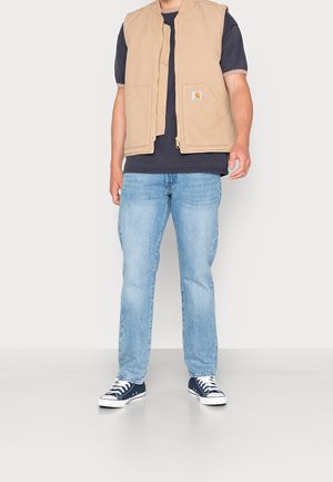 Gilet en coton beige avec une fermeture éclair et deux poches avant, porté par-dessus un t-shirt marine, associé à un jeans bleu clair et des baskets marines.