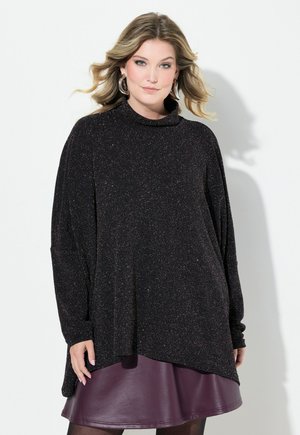 Vrouw met golvend blond haar draagt grote oorringen, een zwarte glinsterende trui met lange mouwen en een korte donkerpaarse leren rok.