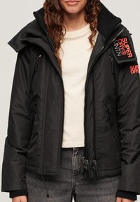 Sort vandtæt jakke med flere lynlåse, høj krave og et mærke med "SUPERDRY" i fed rød. Lavet af struktureret stof.