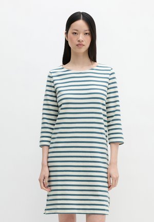 Kobieta stoi ubrana w białą i miętową sukienkę w poziome paski z rękawami do łokcia na jednolitym białym tle.