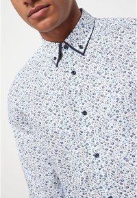 Bílá košile s knoflíky s modrým květinovým vzorem, kontrastním tmavě modrým límcem a knoflíky, vyrobená z lehké látky s hladkou texturou.