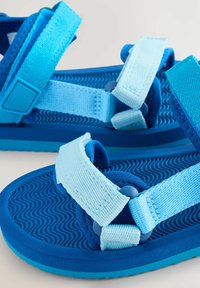 Sandalias azules con suelas de goma texturizadas, correas ajustables en azul claro y oscuro, y acentos de herrajes circulares.