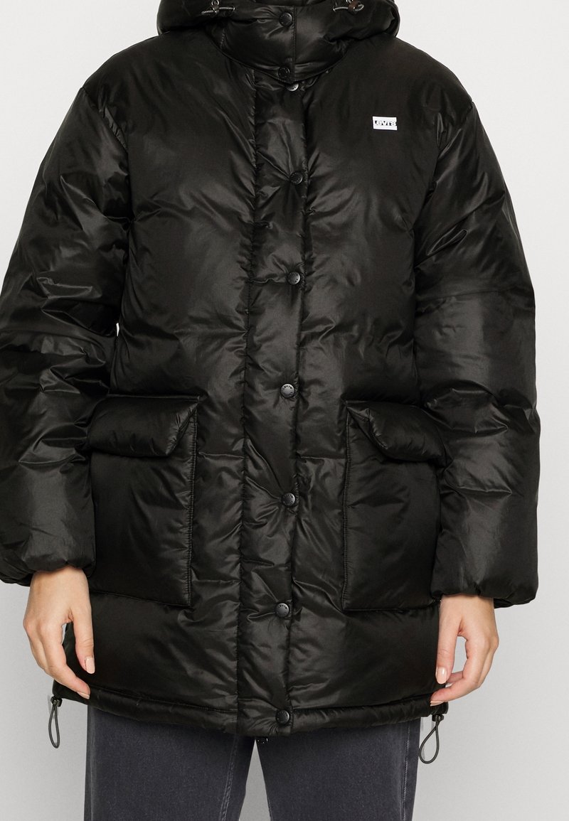 Veste rembourrée noire avec capuche, boutons pression, deux poches avant et cordons de serrage réglables au bas. Matériau brillant et isolé.