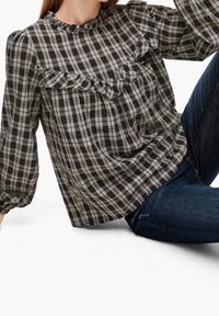 Femme portant un chemisier à carreaux noir et blanc avec un détail à volants, associé à un jean bleu foncé, assise avec un genou relevé.