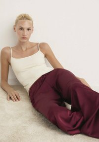Top in crema con spalline sottili abbinato a pantaloni larghi in satin bordeaux. La trama è liscia, in contrasto con il fondo di un tappeto chiaro.