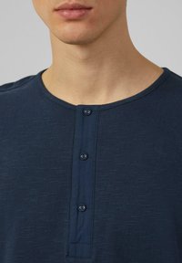 Marineblauw henley-shirt met een ronde hals, drie knoopsluiting en textuurstof. Zichtbare stiksels langs de kraag en knoopsluiting.