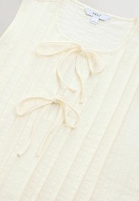 Haut sans manches de couleur crème avec un design texturé à rayures verticales et trois nœuds noués à l'avant.