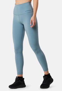 Legging taille haute bleu clair en matériau extensible, présentant une texture lisse et un logo à la taille. Chaussures de sport noires.