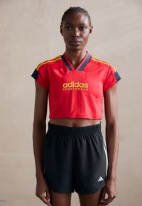 Sarkans īsais sporta krekls ar dzelteno "adidas Sportswear" logotipu un tumši zilām detaļām, kopā ar melniem sporta šortiem ar baltām svītrām.