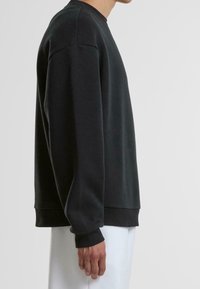 Personne portant un sweat-shirt noir à manches longues avec des poignets et un ourlet côtelés, vue de profil sur un fond uni.