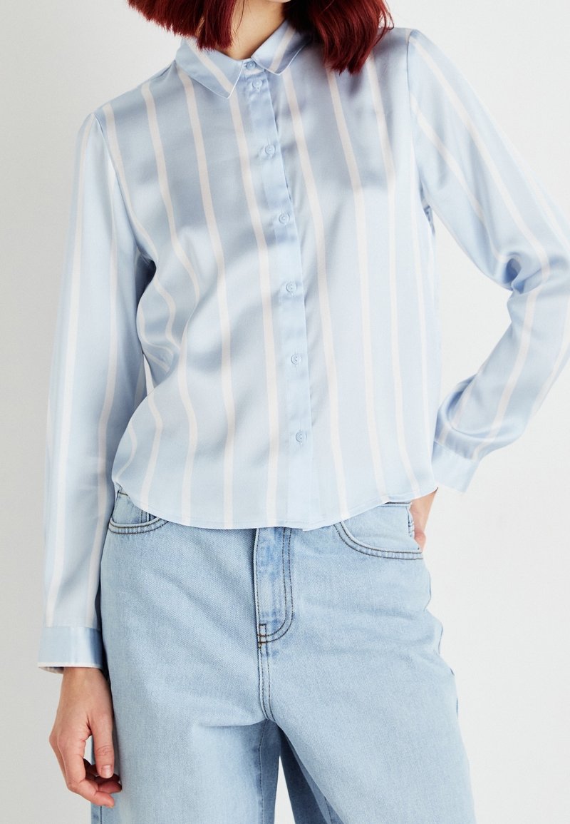 Femme portant une chemise bleu clair à manches longues, rayée verticalement, rentrée dans un jean taille haute bleu clair.
