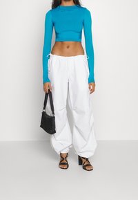 Långärmad turkos avkortad topp, vita byxor med dragsko, svart miniväska och strappriga svarta klackar. Har figursydd design och mjuk textur.