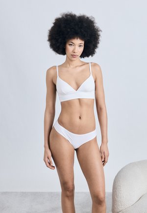 Femme aux cheveux afro portant un soutien-gorge blanc et une culotte, debout près d'un fauteuil blanc texturé sur un fond uni.