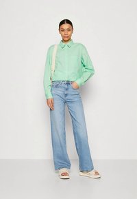 Camisa verde clara abotonada con dos bolsillos, combinada con vaqueros de pierna ancha en azul claro. La modelo lleva una correa crema cruzada en el hombro y zapatos claros.