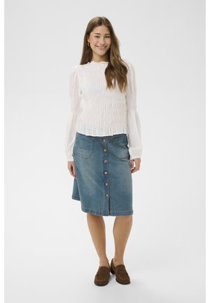 Femme souriante aux cheveux longs portant un chemisier blanc froncé, une jupe en jean boutonnée mi-genoux et des mocassins marron, debout devant un fond uni.