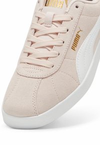 Gros plan sur une chaussure de sport en daim beige Puma avec une bande latérale blanche, des lacets beiges et un logo Puma doré sur la languette et le côté.