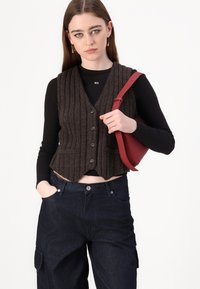 Hnedý rebrovaný pletený crop top s gombíkovým zapínaním, nosený cez čierne tričko s dlhým rukávom. Model drží červenú kabelku na rameno. Tmavé džínsy.