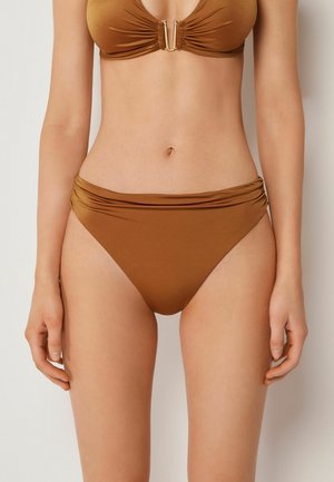 Kvinde iført brune bikiniunderdele med høj talje og en matchende bikinitop med en gylden U-formet spænde i midten.