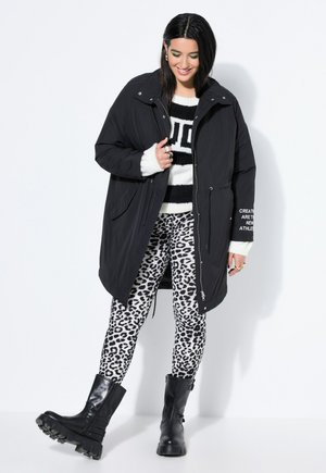 Nainen hymyilee, pukeutuneena mustaan pitkään takkiin, mustavalkoraidalliseen neulepaitaan, pantterikuvioisiin legginsseihin ja mustiin massiivisiin saappaisiin, seisoo valkoisen seinän edessä.