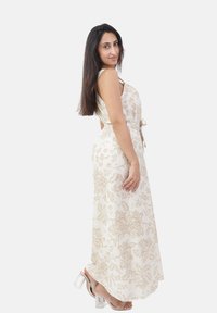 Frau, die seitlich steht, mit langen dunklen Haaren, gekleidet in einem ärmellosen weißen Kleid mit beigen Blumenmustern und klaren High Heels.
