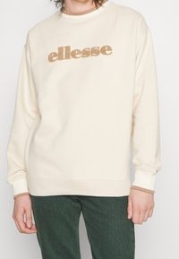 Krämfärgad sweatshirt med en texturerad guld "ellesse"-logotyp. Ribbad avslutning vid ärmar och halsringning, kombinerad med mörkgröna mönstrade byxor. Avslappnad passform.